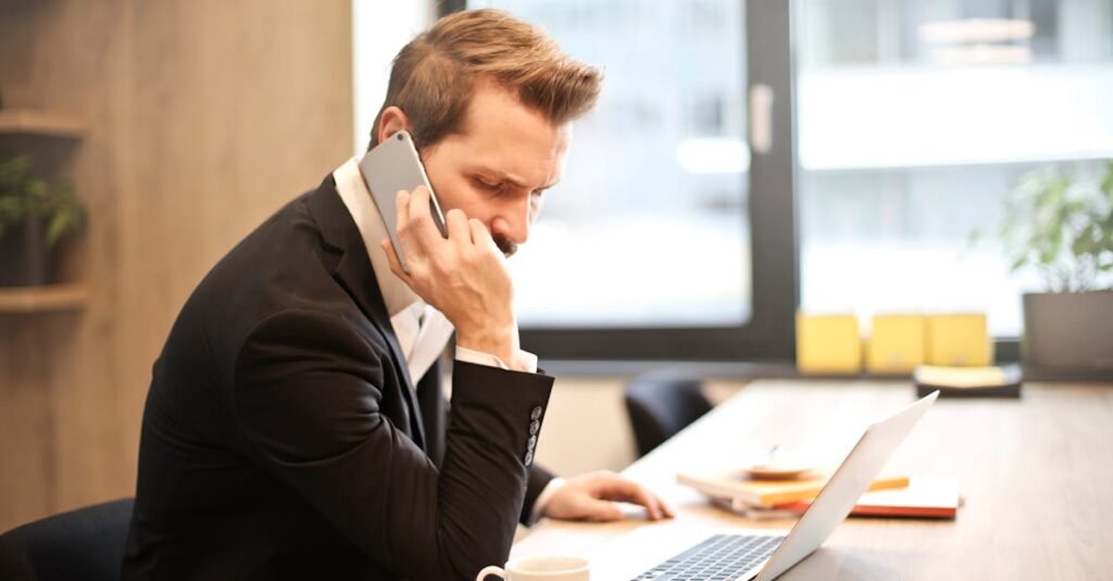 A businessman in a suit making a phone call while working on a laptop in a modern office setting.